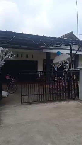 The video pans across a residential street lined with houses and small businesses. Multiple Indonesian flags, red and white, are visible, suggesting a national holiday or celebration. The street is unpaved in some areas, with some debris on the ground. Several motorcycles are parked along the street. In one scene, a small market stall is set up with produce displayed in wooden crates. The overall atmosphere appears to be that of a typical day in a rural or semi-urban Indonesian community.
