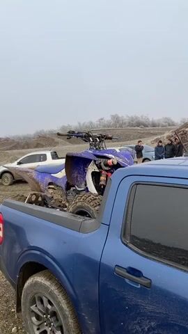 The video captures an outdoor scene, likely in Tolhuin, Argentina, on an overcast and cold day, suggested by the hazy sky and what appears to be frost on distant trees. In the foreground, a blue Ford Maverick pickup truck is parked, with a muddy, blue ATV (all-terrain vehicle) loaded in its bed. Two pairs of shoes are placed on the ground next to the truck.

As the camera pans to the right, a man in a bright green jacket, dark pants, and glasses walks towards the viewer, carrying what appears to be a helmet in his left hand. He is walking away from an older model white pickup truck, which also has a dirty white ATV loaded in its bed. This white truck displays the license plate "EOZ 227" and "4WD" text on its mudflaps. Several other individuals are visible standing near the white truck in the background.

The ground is a mix of dirt and gravel, characteristic of an off-road or quarry environment, with barren hills forming the backdrop. The dirty condition of both ATVs and trucks indicates recent off-road activity. The overall scene suggests a group of people either concluding or taking a break from quad biking in a rugged, outdoor setting.
