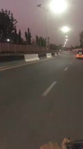 The video shows a first-person perspective of someone riding on a smooth, gray asphalt road. The road is lined with white and black barriers on the left side and is marked with dashed white lines. Tall streetlights illuminate the road. In the distance, there are trees, buildings, and what appears to be a mosque with minarets. A small, yellow motorized vehicle is driving ahead in the same direction. The sky is a dusky pink color, suggesting it is either dawn or dusk. The hand of the person riding is visible in the foreground.