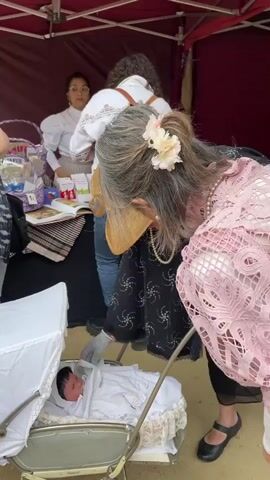 In an outdoor setting, three older women are seen closely examining an antique baby carriage with a dark-haired baby in it. The carriage is off-white with silver-tone metal supports. It is set up in front of a vendor's table, which has a variety of items for sale under a red and white tent. The first woman, wearing a light pink lace top and satin skirt, is positioned on the right of the carriage. The second woman, who stands in front of the carriage, is wearing a straw hat, a pearl necklace, a black dress with a star-shaped pattern, and gray gloves. She has her hands close to the baby. The third woman, who is facing away from the camera, is wearing jeans, black boots, and a white top with embroidery, and a brown backpack. To the left of the carriage, there are other shoppers, a vendor at the table with glasses and a white blouse, and more antique merchandise for sale.