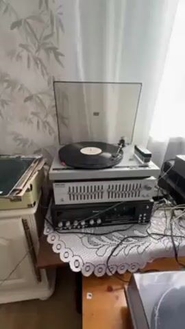 The video shows an audio system set up near a window covered by sheer white curtains. The system includes two turntables, one with a vinyl record playing. Below the operating turntable, there is an equalizer and a receiver. Adjacent to the turntable setup, there's another stack of audio components including an amplifier, and a receiver. Speakers are situated next to this stack. A wooden chest sits on top of one of the speakers. A small antique-style cabinet stands to the left of the audio setup. A lace-edged tablecloth covers the table holding the equipment. The wallpaper behind the setup features a floral pattern. Cables and wires are visible connecting the components.