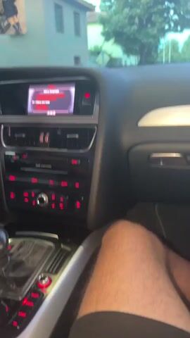 The video is filmed from inside a car, showing the dashboard and a person's leg. The car is an Audi, as indicated by the logo on the screen. The screen displays a menu with various options, possibly related to settings or navigation. The person's leg, which is hairy, is resting on the passenger seat or the center console. Outside the car, trees and buildings are visible, suggesting the car is either parked or moving in a town or suburban area. The lighting suggests it is daytime, possibly late afternoon or early evening, due to the soft, diffused light. The overall impression is a casual, in-car scene.