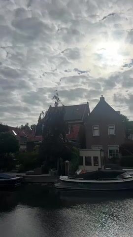 The video shows a canal with two boats docked. On the bank of the canal are several houses, some with red tile roofs and dormer windows. A tall tree with dark foliage stands in front of one of the houses. The sky is mostly cloudy with the sun shining through, creating a bright glow. The water in the canal is dark and reflects the sky and surrounding environment.