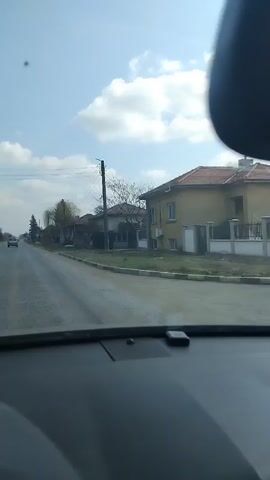 The video shows a view from inside a car driving on a road. The sky is blue with scattered clouds. The road is straight and there is another car driving ahead. On either side of the road are houses and trees. There is a stop sign visible on the right side of the road. The camera is stationary within the car.
