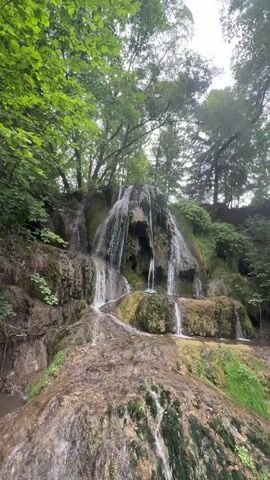 This video shows a multi-tiered waterfall cascading down a moss-covered rock formation. Lush green trees and foliage surround the waterfall, with sunlight filtering through the canopy, creating dappled light on the water and rocks. The water flows into a pool at the bottom of the falls. The scene appears to be a natural, serene environment.