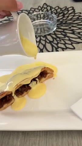 A close-up shot shows a hand pouring a thick, yellow sauce from a white pitcher onto a pastry that is already covered in the same sauce. The pastry, which appears to be a strudel with a visible filling, is on a white rectangular plate. The plate is set on a gray wooden surface. In the background, there is a black placemat with a floral design and a clear glass bowl. The lighting is bright, suggesting an indoor setting, possibly a restaurant. The action focuses on the pouring of the sauce, creating a dynamic visual of the dessert being dressed.