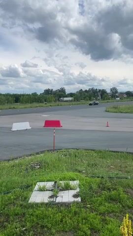 This video captures an outdoor scene at Svetloe Pole, Russia, on a cloudy day. The primary focus is on a black car navigating a temporary driving course marked by orange traffic cones and white and red barriers. The car appears to be engaged in some form of driving exercise, possibly a race or a skill demonstration, given its positioning around the cones.

In the background, several large industrial buildings and a smaller structure with text signage can be seen. The signage on the smaller building reads "КОФЕ & ХОТ-ДОГ," indicating it is a cafe or food stand. A red truck is visible in the distance, and other vehicles are parked in the background. The overall setting is an open, paved area, likely a training ground or a closed-off section of an industrial zone, with a grassy border in the foreground. The weather appears overcast, contributing to a muted lighting condition. No people are clearly discernible within the car or the immediate vicinity of the driving course, but figures can be seen inside the "КОФЕ & ХОТ-ДОГ" structure.
