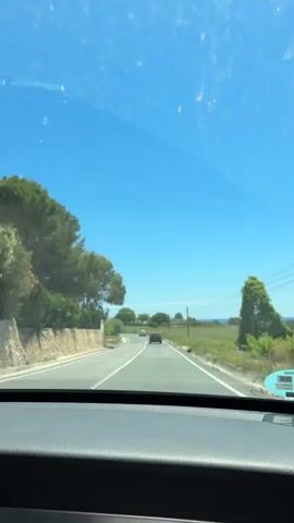 The video shows a driver's point of view while driving on a rural road during a sunny day. The road has a single lane in each direction with a solid white line separating them. There are green trees and shrubbery lining the sides of the road.  Several cars are visible in the distance. A few telephone poles are visible in the distance. The dashboard of the car is visible in the lower portion of the video. A bright blue sky is seen through the windshield. There is music playing in the background.