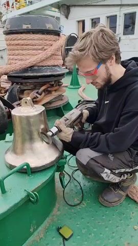 A young man crouches on the green deck of a ship, meticulously polishing a brass bell with a power drill. He wears safety glasses, work gloves, and durable work boots as he focuses on his task. A coil of rope sits nearby on a large winch. The weather is overcast, casting a neutral light on the scene. The man's concentration is evident as he moves the drill carefully across the surface of the bell.