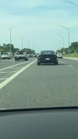 The video shows a black Dodge Charger driving on a highway during the day. The camera is positioned inside another vehicle following the Charger. Other vehicles, including a white SUV and a utility truck, are visible in the surrounding lanes. The highway has several lanes in each direction and is lined with light poles and green exit signs. The sky is partly cloudy. The Charger has a license plate that reads "B2761 EL". The brake lights illuminate briefly.