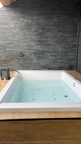 The video shows a close-up of a white bathtub filled with clear, bubbling water. The bathtub is set into a wooden platform and is surrounded by a dark gray stacked stone wall. A silver faucet is visible on the wall behind the tub, and a small black object, possibly a container, sits on the left side of the tub. The water is being poured into the tub from the faucet, creating ripples and bubbles. The overall impression is one of a luxurious and relaxing spa-like environment.