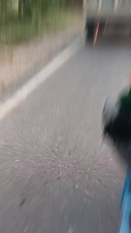 The video shows a point-of-view shot from a vehicle moving on a paved road. The road is gray with a white line on the left side. On the right side of the road, there are trees and foliage. In front of the vehicle, a blue tractor is visible with people in the back. The tractor has colorful tassels hanging from the top. The words "BLOW" and "HORN" are painted on the back of the tractor in large letters. The camera is moving, creating a blurred effect.