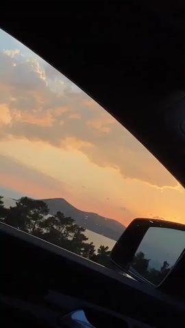 The video shows a view from inside a car looking out the window at a sunset over a body of water and mountains. The sky is filled with colorful clouds in shades of orange, pink, and white. The sun is low on the horizon, casting a warm glow. Trees and a side mirror are visible in the foreground.
