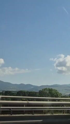 A POV shot from inside a vehicle shows a sunny, clear sky with scattered clouds. The view is of a rural landscape with rolling hills and mountains in the distance. In the foreground, there is a metal guardrail and a road. As the vehicle moves, a billboard and a power line tower come into view. A car passes by the power line tower.