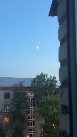 The video captures a view from a window at dusk. The sky is a clear, pale blue, transitioning from day to night. Two celestial bodies are visible in the sky; one is a crescent moon, illuminated on its right side, and the other appears to be a smaller, fainter object positioned above and slightly to the left of the moon. In the middle ground, a building with multiple floors is visible. It has a brick facade with numerous windows, some of which are lit from within, suggesting occupancy. A metal roof is visible on top of the building. Several trees with green foliage are scattered in front of and around the building. The foreground of the video is partially obscured by a rough, textured surface, possibly a brick wall or the window frame itself, which is out of focus and takes up the right side of the frame. The overall impression is a quiet, urban scene at twilight.