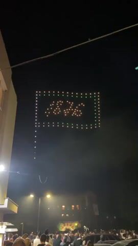 At night, a crowd of people stands on a street, looking up at a drone display in the sky. The display is shaped like a flag with a rectangle containing a symbol made of lights. The rectangle's border consists of rows of white, green, and yellow lights. Below the drone display, streetlights illuminate buildings and the crowd. A building is visible on the left of the frame. Power lines extend across the top of the scene.