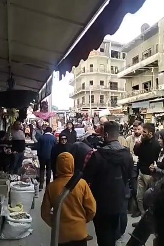 The video shows a crowded outdoor market, likely in the Middle East. Several people are walking through the market, including a group gathered around what appears to be a film crew. The crew is clustered around an older man with glasses and a red and black plaid shirt, possibly directing him. There are market stalls and shops on either side of the main walkway. In the background, a tall, multi-story building with an architectural style common to the region is visible.