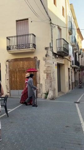 A street scene in front of a building with a large wooden door and balconies. Two people are walking down the street. The person on the left is wearing a long, red, ornate dress and a hat with a red umbrella attached. The person on the right is wearing a gray suit and holding a cane. In the background, other people are walking down the street, also dressed in historical clothing. The street is paved with cobblestones and there are buildings on both sides. The lighting suggests it is late afternoon or early evening.