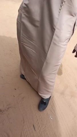 Two men are standing outdoors on a dirt ground. One of the men is wearing a long, light tan robe, black shoes, a white beaded necklace, sunglasses, and a small, patterned hat. The other man is wearing a green robe and black sandals. The man in the tan robe has his arm around the other man's shoulder. The man in the tan robe points at something off-camera. The man in the tan robe then walks away. There are trees and buildings visible in the background.