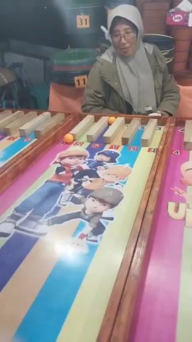 A woman wearing glasses and a hijab is playing a carnival game. She is seated behind a long wooden table with several lanes, each containing wooden obstacles and small orange balls. The lanes are decorated with colorful cartoon characters. The woman is shown leaning forward, looking at the game, and her hands are positioned as if she is playing. In the background, there are some barrels with numbers on them and other carnival stall elements.
