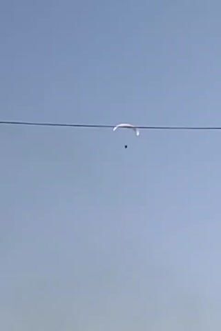 The video shows a group of people wearing helmets and black shirts gathered on a street with buildings on either side. Above them, a paraglider with a box suspended underneath is visible against a clear sky. There is smoke rising in the distance behind the buildings.