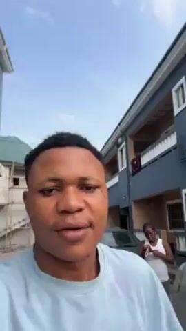A young man is filming a selfie video while standing in a driveway.  He is smiling at the camera. In the background, a woman in a white tank top and gray sweatpants is standing in front of a gray car, appearing to look at something in her hands. Behind the woman, there is a two-story blue building with white-framed windows. To the left of the man, there is a smaller, partially-constructed building with scaffolding around it. The sky is overcast.