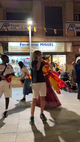 A young male with blonde hair, wearing a black t-shirt and white shorts, stands in the middle of a street at night. He is holding a Spanish flag in his right hand and a red object, possibly a phone, in his left hand, bringing it to his ear as if making a call. He is looking to his left. Behind him is a store with a sign that reads "Ferreterías MIGUEL". In the background, other people are visible walking and standing around. The street is paved and illuminated by streetlights.