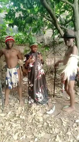 Three children dressed in costume stand in a wooded area covered in fallen leaves. The child on the left wears a red and white striped hat, a patterned cloth around their waist, and holds a cane. The child in the center wears a long dress with a floral pattern and a green hat. The child on the right wears a brown hat, a patterned cloth around their waist, and holds a cane. All three children have dark makeup on their faces. They are standing still and looking at the camera.