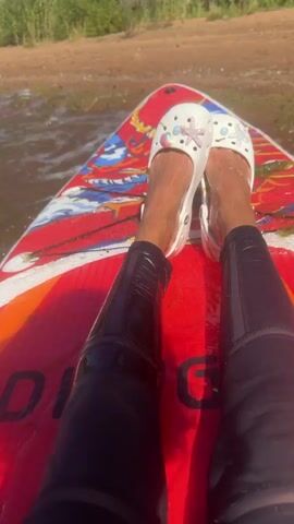 The video shows a person's legs and feet from a first-person perspective while they are on a paddleboard in the water. The person is wearing black leggings that are wet and white Crocs with colorful charms. The paddleboard is red with a colorful design. The water is choppy, and there are reeds or tall grass in the background on the shore. The sun is shining, creating reflections on the water and the paddleboard. The person appears to be relaxed and enjoying the activity.