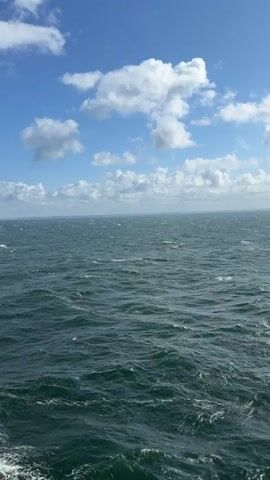A vertical video captures a wide expanse of the ocean under a partly cloudy sky. The water is a deep teal color and appears choppy with small to medium-sized waves. Whitecaps are visible on some of the waves, indicating a moderate breeze. The horizon line is visible in the distance, and the sky above is a bright blue with scattered cumulus clouds that are white and fluffy. The camera perspective is from a high vantage point, possibly from a ship, looking out over the water. The lighting suggests it is daytime, with sunlight illuminating the clouds and the surface of the water. No people or objects are clearly visible in the frame, focusing solely on the natural environment of the sea and sky.