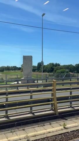 The video shows a view from a moving vehicle, likely a train, looking out a window. A large, rectangular monument with inscriptions and decorative carvings is visible in the distance, set against a backdrop of green trees and a clear blue sky. In the foreground, there is a yellow metal railing and structure, suggesting the vehicle is on an elevated track or bridge. The monument appears to be made of light-colored stone and has a weathered appearance. The surrounding environment is open and green, with some trees and sparse vegetation. The video pans slightly to the right, showing more of the railing and the landscape.