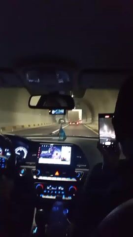 The video shows the interior of a car driving through a tunnel at night. The driver's side dashboard with a speedometer and infotainment screen is visible on the left. A passenger in the right seat is holding a smartphone and recording the scene. The infotainment screen displays a video of a cartoon character. There are other vehicles, including trucks, visible ahead in the tunnel. The tunnel walls are lit and appear to be made of concrete. The lighting inside the car is primarily from the dashboard and the phone screen, with blue ambient lighting visible.