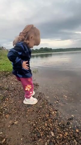 A young child with red hair, wearing a dark jacket with a blue and black pattern and pink pants with floral designs, stands at the edge of a body of water. The child is looking down at the water and the pebbly shore. The child then turns to look at the camera and takes a step backwards. The video then pans to show the expanse of the water and the cloudy sky. The environment appears to be a natural outdoor setting, possibly a lake or river, with trees visible in the distance.