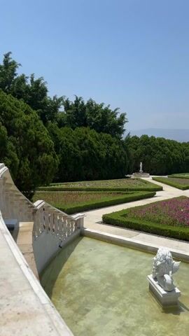 The video shows a beautiful, manicured garden with a large fountain at the center. The garden is laid out in a geometric pattern with flower beds edged by trimmed hedges. Walkways intersect the flower beds, and several people are strolling through the garden. In the foreground is a pool of water with white lion statues. Beyond the garden, trees line the horizon under a clear blue sky.