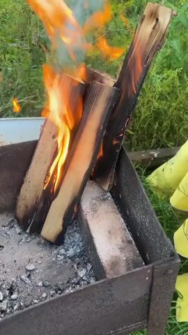 A close-up video shows three pieces of wood burning in a grill. The wood is oriented vertically, with flames licking up the sides. Ash and embers are visible at the bottom of the grill. The background is blurred and appears to be an outdoor setting with green foliage. A person's yellow-gloved hand is visible on the right side of the frame.