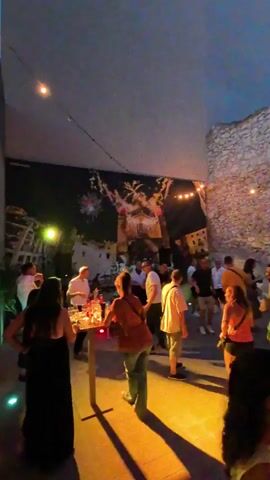 The scene is set at an outdoor event, likely a party or gathering, occurring at dusk. There are approximately 20 people present, many of whom are standing or moving around.  The backdrop features a large mural of a building or town illuminated by fireworks, suggesting a celebratory theme. String lights are hung overhead, contributing to the ambience. A small bar table, with various bottles and glasses on it, is centrally located, where some people are congregating. One wall of the space is made of stonework, adding texture and visual interest to the environment. The people are dressed in casual attire, and the overall atmosphere appears relaxed and social.