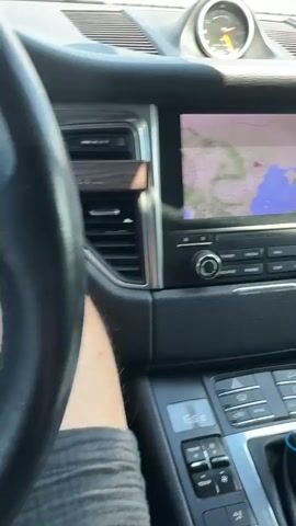 A first-person perspective video shows the interior of a car. The camera pans from the center console to the steering wheel. In the center console, a navigation screen displays a map, and below it are climate control and audio system buttons. The gear shifter is visible in the foreground. The camera then moves to the steering wheel, revealing the Porsche logo and the car's dashboard gauges. The car appears to be parked, as indicated by the gauges and the absence of movement. The overall lighting suggests it is daytime.