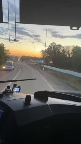 The video shows the interior of a bus cab as it drives down a road at sunset. The driver's hands are visible on the steering wheel. The dashboard displays various screens, one of which shows a navigation map. Outside the windshield, the sun is low on the horizon, casting a warm glow over the landscape. Trees line the sides of the road, and streetlights are visible in the distance. A road sign indicating a sharp curve is also visible. The sky is filled with wispy clouds.