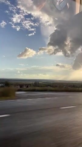 The video shows a view from a moving vehicle looking out at a rural landscape. The road is wet and reflects the sky. There are clouds in the sky, with sunlight breaking through them. The landscape includes fields, trees, and some buildings. The video is shot at an angle, giving a sense of motion.