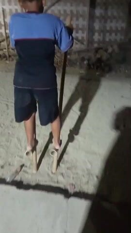 A young person stands on stilts made of bamboo, holding onto two long poles for balance. They are outdoors on a sandy surface, with a partially constructed building visible in the background. The person is wearing a blue and black t-shirt and black shorts. The scene appears to be at dusk or night, with artificial lights casting long shadows.