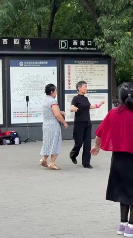 The video captures an outdoor scene featuring three women in what appears to be a public square or plaza, with a Beijing subway station entrance visible in the background.

Two women are actively engaged in a partner dance. The woman on the left is dressed in a light blue patterned dress, resembling a modern cheongsam, and gold low-heeled shoes. Her dance partner, positioned with her back mostly to the camera, wears a black short-sleeved top, black wide-leg pants, and black shoes. They move gracefully, holding hands and performing coordinated steps, suggesting a ballroom or square dancing style.

Observing the dancers from the foreground, facing away from the camera, is a third woman. She is distinctive in a bright red short cape with an intricate pattern, worn over a black dress or skirt, paired with black sheer leggings and vibrant purple sneakers with white soles. Her hair is tied back in a ponytail.

Behind the dancers, a prominent black sign displays "D 西南口 South-West Entrance" in white characters, clearly identifying a subway station. To its left, another large poster shows a "北京城市轨道交通线网图" (Beijing Urban Rail Transit Network Map), indicating the location is in Beijing. A tripod holding a smartphone or camera is set up near the map, likely recording the dance. Further background details include green trees and foliage, suggesting a park-like setting, and other indistinct figures along with parked bicycles.

The lighting indicates it is daytime, possibly late afternoon or early evening, with clear weather. The scene portrays a casual moment of public recreation and community activity.