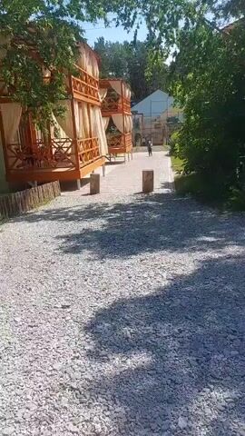 The video shows a gravel path leading towards a paved walkway lined with trees. On the left side of the walkway are two-story wooden structures with balconies draped in sheer white curtains. These structures appear to be cabanas or guest rooms. A person is walking away from the camera on the paved walkway. In the background, there are more buildings and dense green trees under a bright, sunny sky. The overall setting suggests an outdoor resort or lodging area.