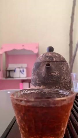 The video shows a close-up of a person's nose and mouth as they lean in to smell a glass of dark amber liquid, likely tea. In the background, a textured, dark-colored teapot sits on a wooden surface. Behind the teapot, a light pink shelf with decorative elements is visible, along with a blurry light-colored wall and a branch. The lighting is soft and diffused. The action is centered around the sensory experience of smelling the beverage.