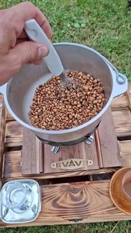 A person's hand holds a whisk, stirring brown beans in a pot. The pot rests on a wooden stand with copper accents and a logo that reads "EVAVE". Beyond the stand, green grass is visible, with a pathway leading into the distance. A small Romanian flag is planted in the grass to the left of the frame. Further in the background, two helicopters and a biplane are displayed on a grassy field, with people walking on a pathway between them. The sky is overcast.