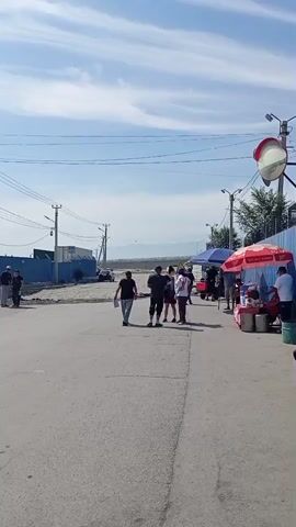 The video shows an outdoor scene with several people. In the foreground, a paved area leads to a sidewalk. On the right side of the sidewalk, there are several stalls with umbrellas providing shade. People are sitting and standing near these stalls. One stall has a red and white umbrella and appears to be selling drinks, with several large containers visible. Another stall to its right has a blue umbrella. Further down the sidewalk, there are more people and stalls. In the middle ground, a group of young men are gathered on the paved area, some appearing to be talking. To the left, there is a street with a vehicle and some construction or industrial fencing. In the background, there are power lines, trees, and distant mountains under a blue sky with scattered clouds. The overall impression is of a public area, possibly a market or a place of gathering, with vendors and customers.