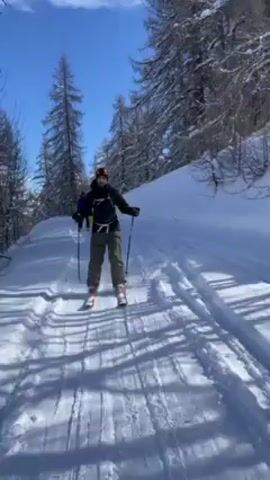 A person is skiing on a snowy path through a forest on a sunny day. The skier wears a black jacket, grey pants, and a black backpack. The path is lined with snow-covered trees. The camera shows the skier's point of view as they glide along the track. The skier appears to lose balance and falls, causing the camera to briefly show the ground.