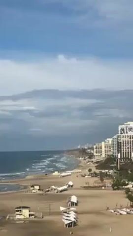 The video shows a high-angle view of a beach on a cloudy day. The beach is sandy and mostly empty, with rows of white sun shelters. There are a few lifeguard stations visible, and some people are walking along the shore. The ocean is rough with white-capped waves. In the background, there is a line of high-rise buildings along the coastline. The sky is mostly covered with gray clouds, with patches of blue visible.