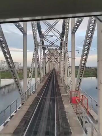 This video shows a first-person perspective of a train crossing a large metal truss bridge over a body of water. The bridge has multiple tracks and is supported by a complex network of steel beams. On either side of the tracks, there are metal railings and some red safety equipment, possibly a fire extinguisher box. Beyond the bridge, the landscape appears to be flat, with grassy fields and some distant trees or structures under a cloudy sky. The camera is positioned inside what appears to be the train's cabin, looking forward along the tracks. The video captures the movement of the train as it traverses the bridge.