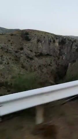 The video shows a passing view of a rocky, mountainous landscape from a moving vehicle. The vehicle is traveling along a road bordered by a white and black guardrail. The terrain consists of dry, scrub-covered hills with rugged rock formations and a deep ravine. In the foreground, vegetation is sparse. Towards the end of the clip, a road sign with red chevrons indicating a sharp turn to the right becomes visible. The sky is overcast and the lighting suggests it is either early morning or late evening. There are no people or animals visible in the video.
