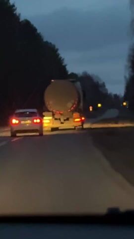 The video captures a forward-facing view from inside a moving vehicle driving on a two-lane road during twilight or early night. The sky is dark and overcast, creating a low-light environment. Dense, dark trees flank both sides of the road, suggesting a rural or forested setting.

Ahead of the camera vehicle, two other vehicles are visible. To the left is a white or light-colored passenger car, possibly a sedan or hatchback, with its red taillights and amber running lights illuminated. To the right, driving in the adjacent lane, is a large dark-colored tanker truck, identifiable by its prominent cylindrical tank. The truck's rear features bright red taillights, several amber marker lights, and reflective red and white chevron-patterned markings on its bumper.

The asphalt road surface appears somewhat damp, reflecting the vehicle lights. In the distance, faint, indistinct lights can be seen further down the road. A prominent and continuous "whoop whoop" siren sound is audible throughout the clip, indicating the presence of an emergency vehicle nearby, although no such vehicle is directly visible within the frame. The scene portrays a routine drive on a road through a wooded area. No discernible text is visible on any of the vehicles or roadside elements.